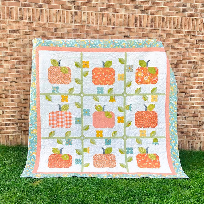 A handmade quilt featuring a grid pattern of orange and red apples with green leaves on a white background, bordered in peach and floral print, displayed outdoors on green grass against a brick wall.
Photo by My Sew Quilty Life [additional_image_link]