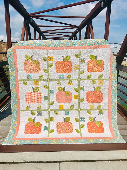 A handmade quilted patchwork blanket featuring orange patterned pumpkins, green leaves, and yellow flowers, displayed outdoors on a wooden bridge.
Photo by My Sew Quilty Life [additional_image_link]