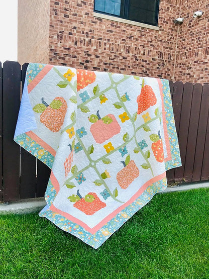 A quilted patchwork blanket featuring strawberries and apples on a vine, draped over a dark brown wooden fence in an outdoor backyard setting with green grass and a brick wall in the background.
Photo by My Sew Quilty Life [additional_image_link]