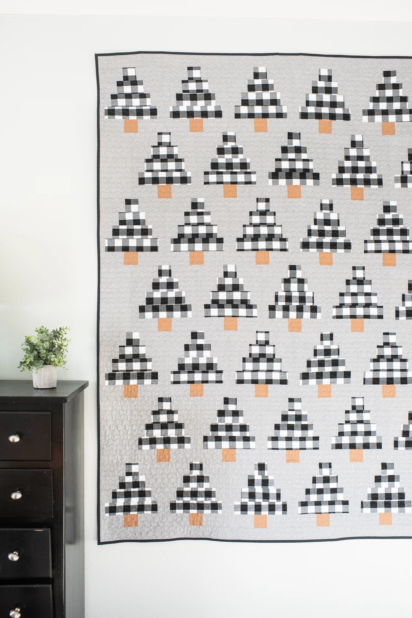 A large quilted wall hanging or blanket with a repeating black-and-white buffalo plaid tree motif and small orange accents, displayed against a white wall in a room with a dark wood dresser and small plant visible on the left.
Photo by Quilty Love [additional_image_link]