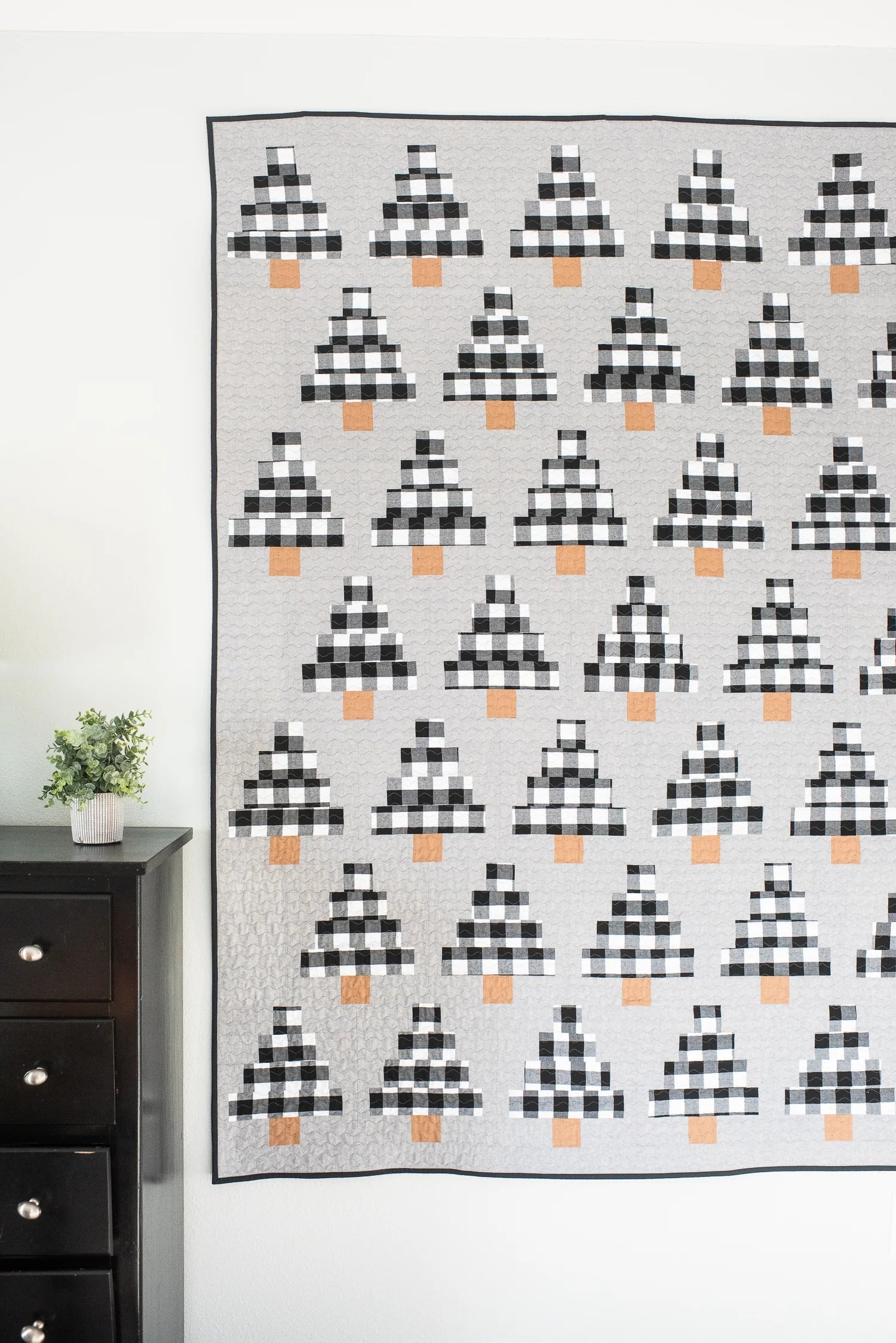 A large quilted wall hanging or blanket with a repeating black-and-white buffalo plaid tree motif and small orange accents, displayed against a white wall in a room with a dark wood dresser and small plant visible on the left.
Photo by Quilty Love [additional_image_link]