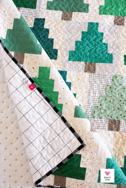 A quilted patchwork blanket featuring green and white geometric Christmas tree patterns, with one corner folded over to show the back side with a black-and-white grid pattern and a small red brand tag. A 'Quilty Love' logo is visible in the bottom right corner.
Photo by Quilty Love [additional_image_link]