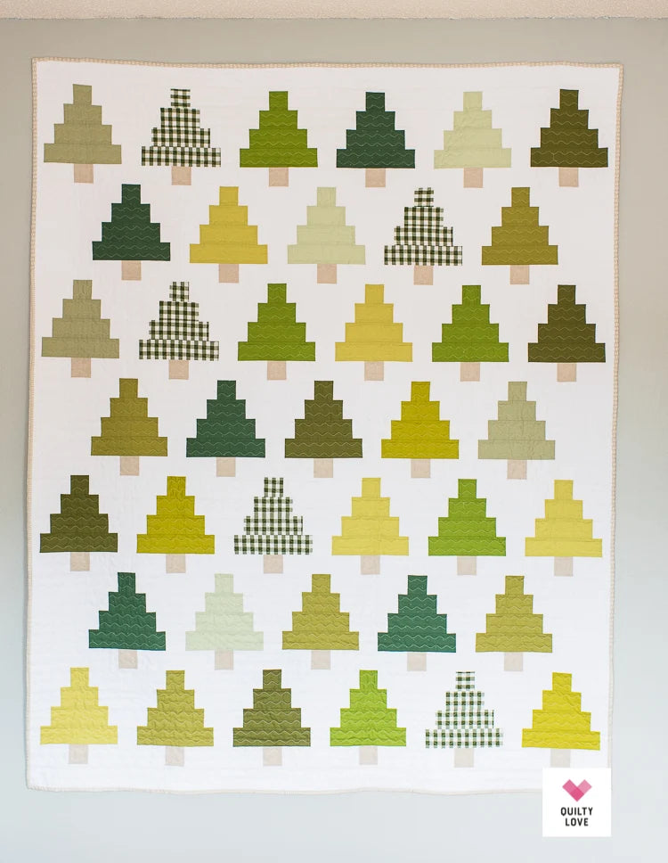 A quilted wall hanging or blanket featuring rows of stylized Christmas tree shapes in various shades of green, yellow, and beige on a white background, photographed against a light neutral wall.
Photo by Quilty Love [additional_image_link]