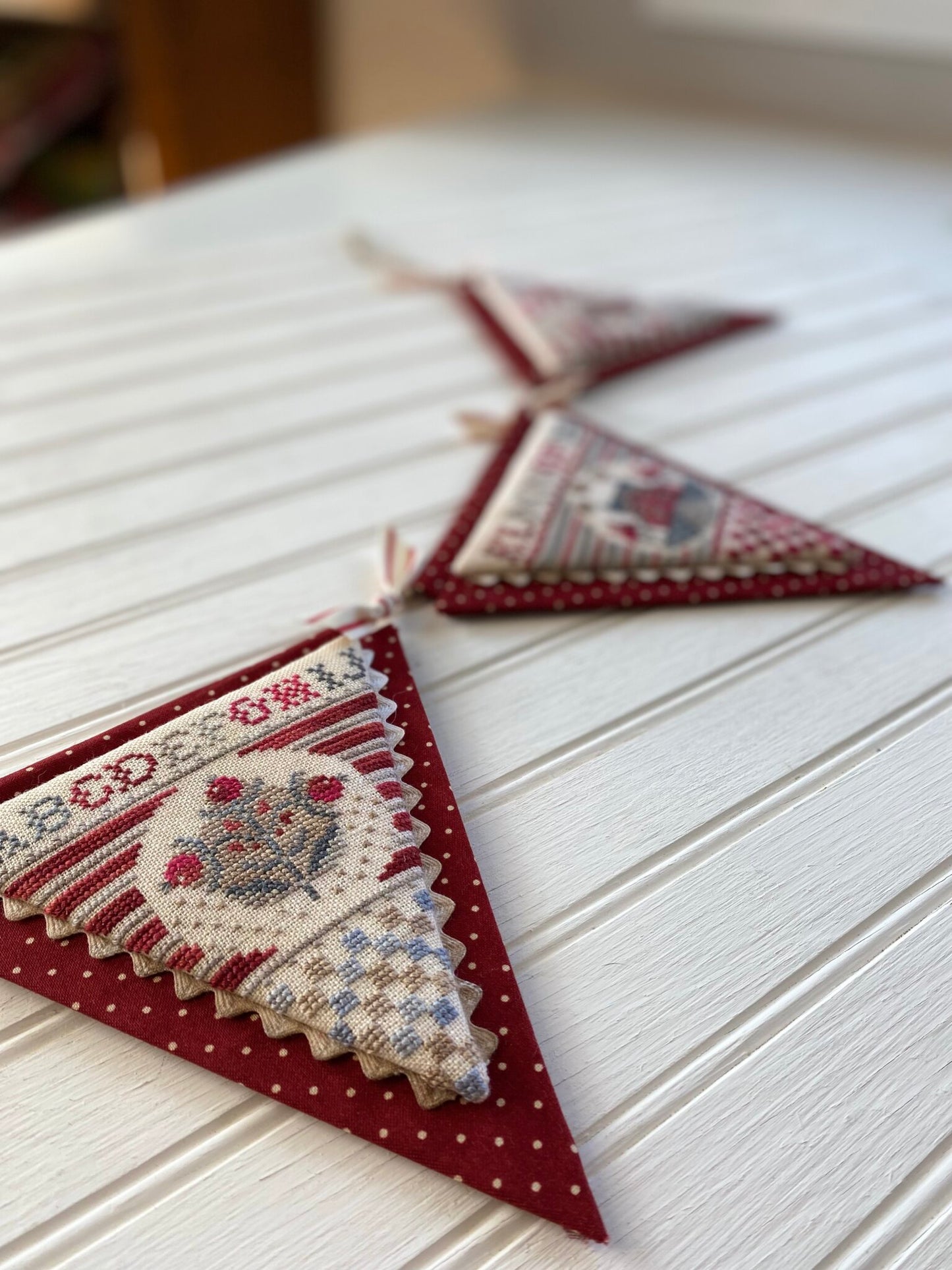 Three triangular fabric bunting flags with red and cream cross-stitch style holiday designs, hanging on a white paneled surface.
Photo by Hands on Design [additional_image_link]