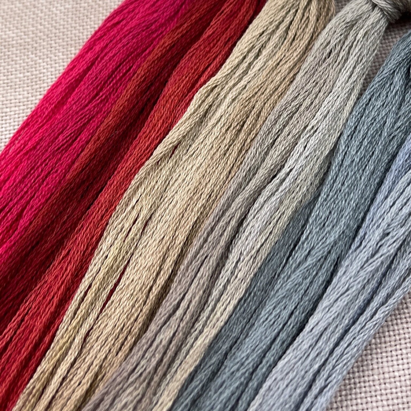 Four skeins of embroidery floss in vertical stripes of bright pink, rust red, warm beige, and dusty blue, laid diagonally on a textured beige fabric background.
Photo by Hands on Design [additional_image_link]
