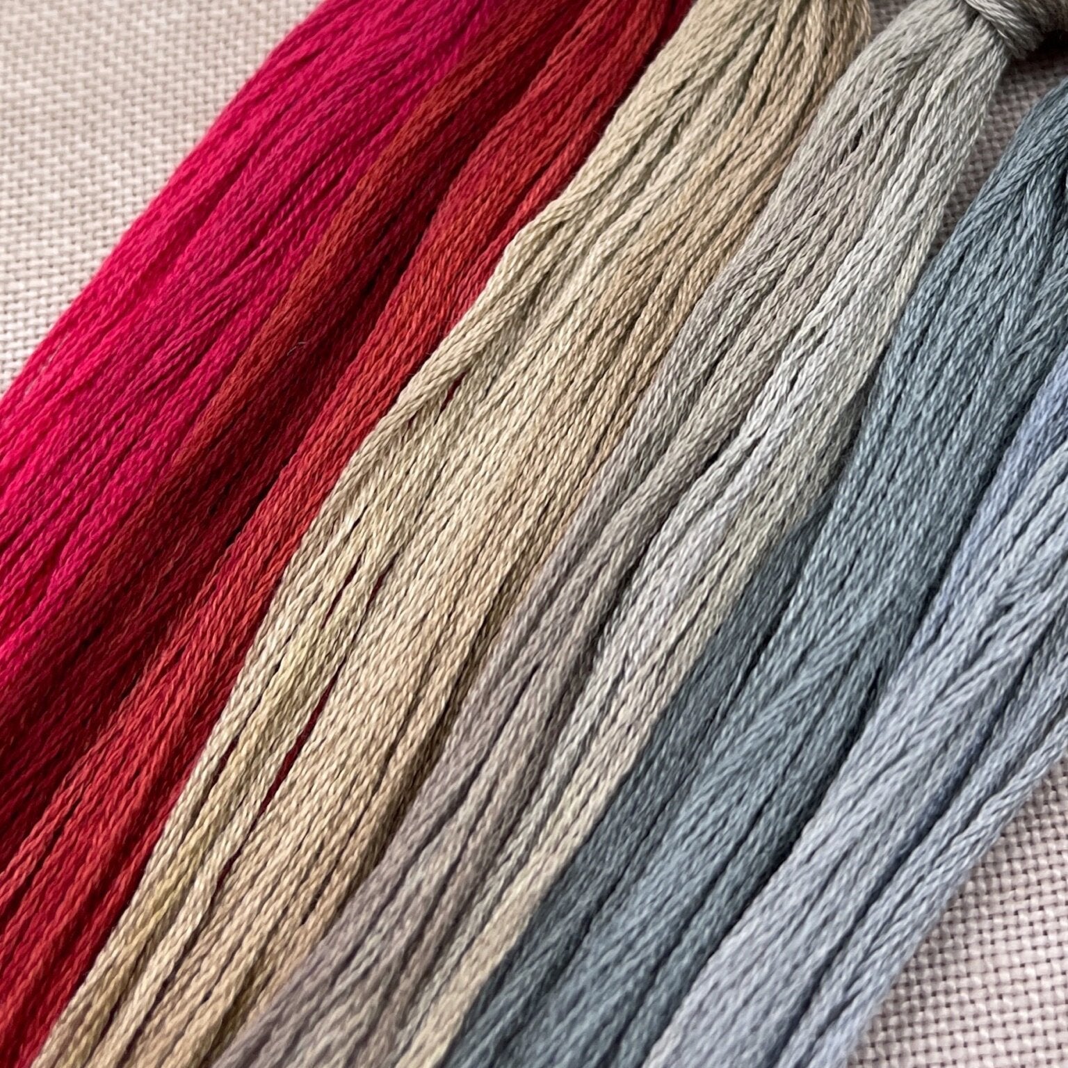Four skeins of embroidery floss in vertical stripes of bright pink, rust red, warm beige, and dusty blue, laid diagonally on a textured beige fabric background.
Photo by Hands on Design [additional_image_link]