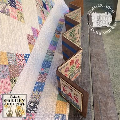 A close-up of a handmade quilt with a zigzag wooden display stand holding fabric panels. The quilt features a patchwork design with various floral and geometric patterns in blue, pink, yellow, and white.
Photo by Summer House Stitche Workes [additional_image_link]