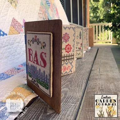 A wooden cross-stitch sampler box standing upright on a rustic wooden table, with a quilt and outdoor porch visible in the background. A 'Lady's Garden Journal' logo is shown in the bottom right corner.
Photo by Summer House Stitche Workes [additional_image_link]