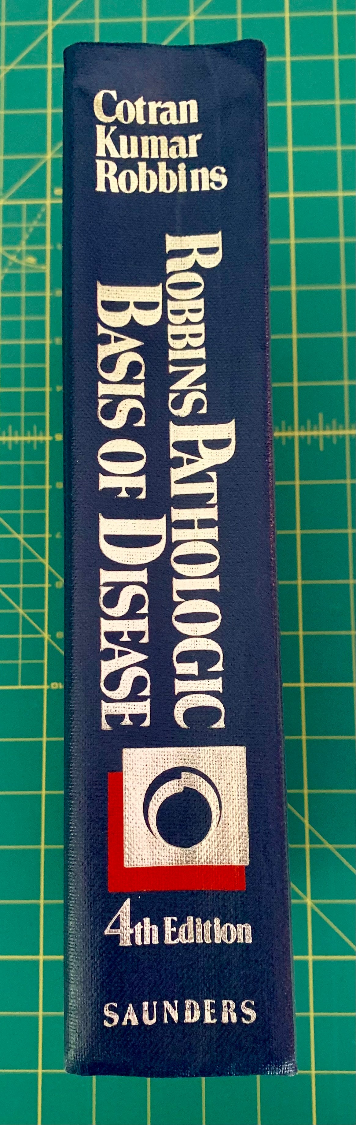 A single book standing upright on a green cutting mat. The book cover is dark blue with large white text reading 'Robbins Pathologic Basis of Disease' and '4th Edition,' plus the Saunders publisher mark near the bottom.
[additional_image_link]