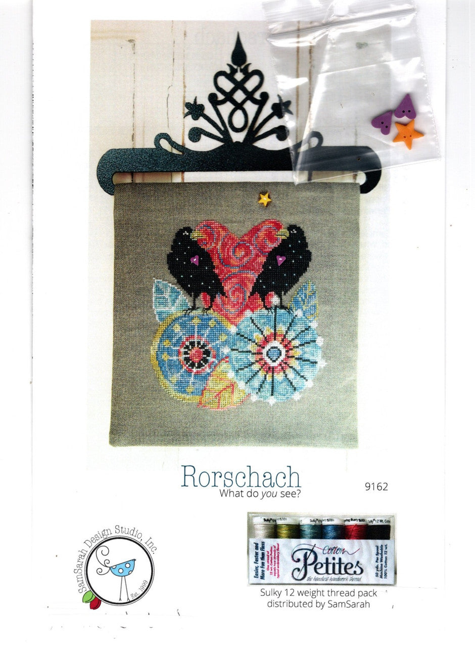 A product display card for the Rorschach 'What do you see?' embroidery pattern, showing a finished multi-colored owl embroidery on linen fabric, a black metal hoop, a small plastic bag of star embellishments, and a Rorschach thread pack at the bottom.
Photo by SamSarah Studio Designs [additional_image_link]