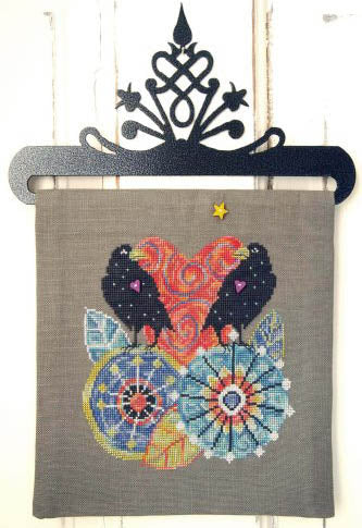 A needlepoint canvas featuring a colorful design of two black birds, a red and orange patterned heart, and two blue and yellow mandalas on a gray linen background, hanging from a black metal rack against a white wood plank wall.
Photo by SamSarah Studio Designs [additional_image_link]