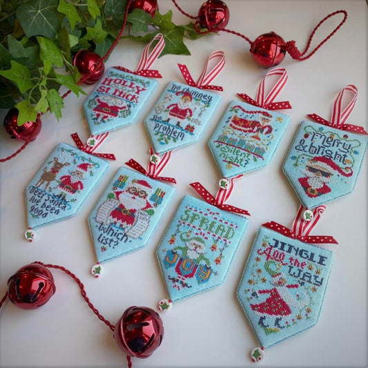 A set of light blue embroidered Christmas pennant tags with red and white striped hanging ribbons, decorated with festive stitched designs and holiday phrases. The tags are arranged on a white surface with red jingle bells and green ivy as styling props.
Photo by HandsOnDesign.biz [image_link]