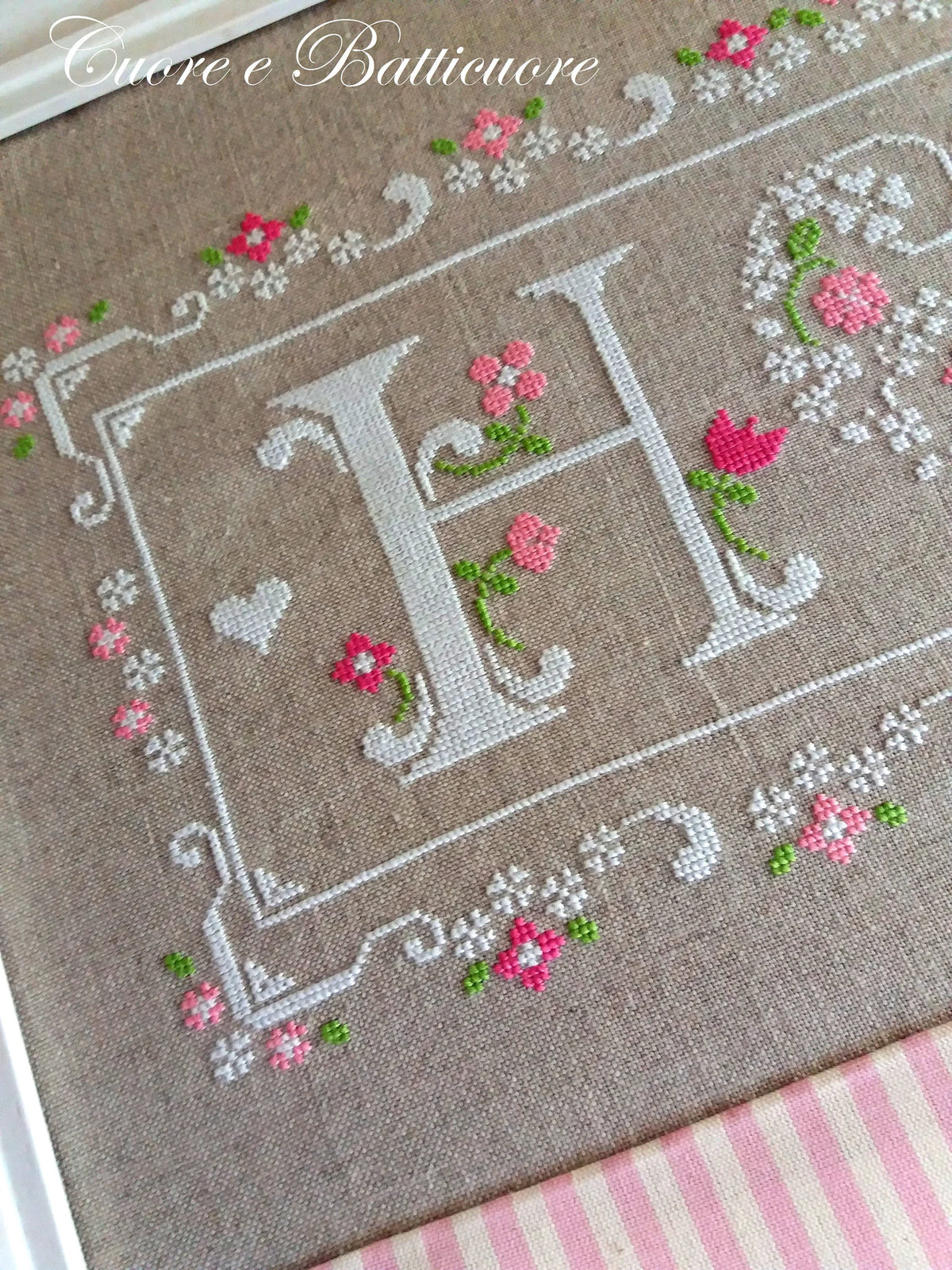 A close-up of a framed cross-stitch piece featuring a large embroidered letter H surrounded by pink flowers and green stems on a beige linen background, with a pink and white striped fabric visible at the bottom.
Photo by CuoreBatticuore.com [additional_image_link]
