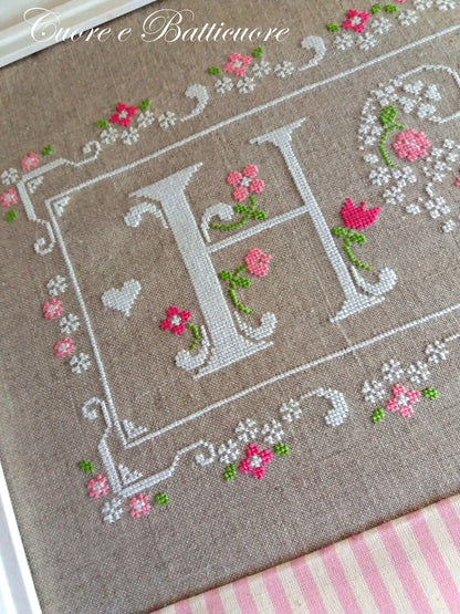 A close-up of a framed cross-stitch piece featuring a large embroidered letter H surrounded by pink flowers and green stems on a beige linen background, with a pink and white striped fabric visible at the bottom.
Photo by CuoreBatticuore.com [additional_image_link]