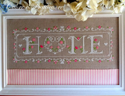 A framed cross-stitch piece featuring the word 'HOME' with 'O' formed by two hearts and floral details. The top half is tan linen with pink flowers and green stems, and the bottom half has pink and white vertical stripes.
Photo by CuoreBatticuore.com [image_link]