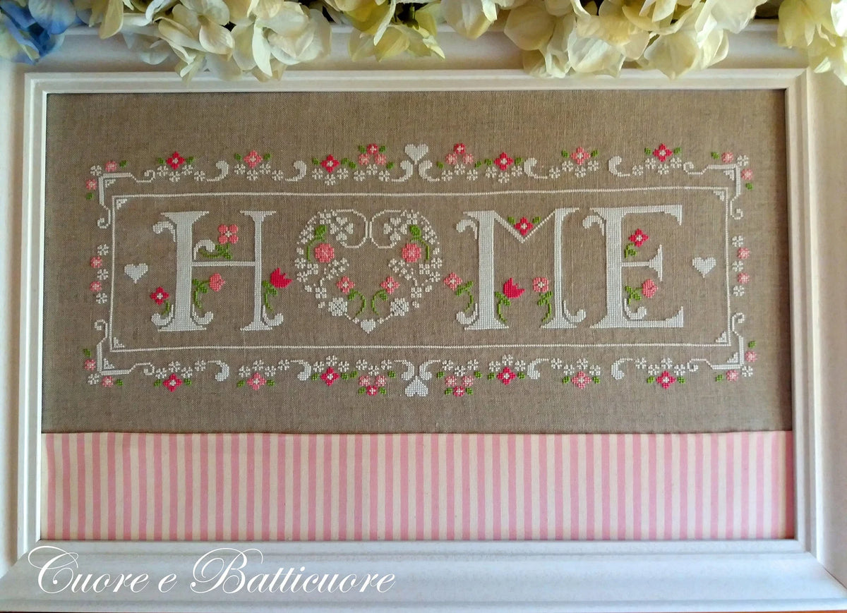 A framed cross-stitch piece featuring the word 'HOME' in white embroidered letters, with 'O' formed by a floral heart design and pink flowers throughout. The top section is tan linen with a pink and white striped band across the bottom, and the piece is displayed in a white frame with decorative flowers above.
Photo by CuoreBatticuore.com [additional_image_link]