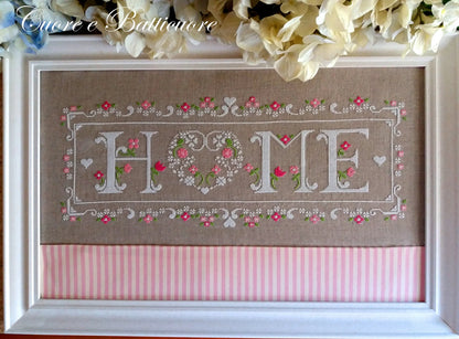 A framed cross-stitch piece featuring the word 'HOME' with a heart-shaped 'O' and floral border details, mounted on a brown linen background with a pink striped bottom panel. The frame is white, and decorative flowers are draped above it.
Photo by CuoreBatticuore.com [additional_image_link]
