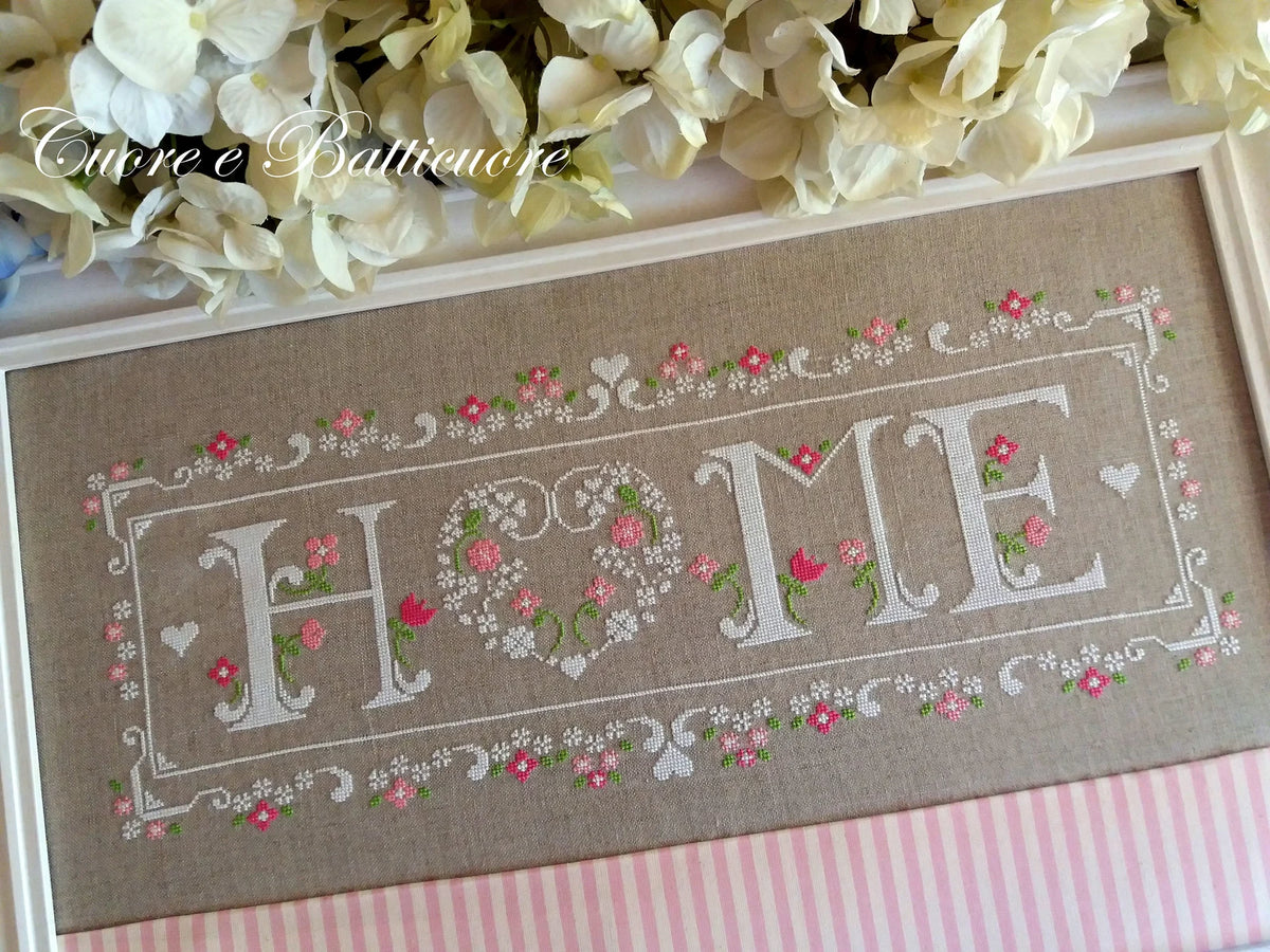 A framed cross-stitch piece featuring the word 'HOME' in white letters with pink and green floral details on a beige linen background, styled with white flowers and a pink striped fabric underneath.
Photo by CuoreBatticuore.com [additional_image_link]