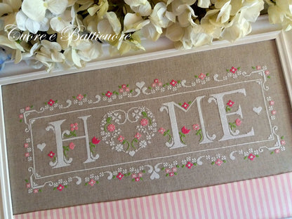 A framed cross-stitch piece featuring the word 'HOME' in white letters with pink and green floral details on a beige linen background, styled with white flowers and a pink striped fabric underneath.
Photo by CuoreBatticuore.com [additional_image_link]