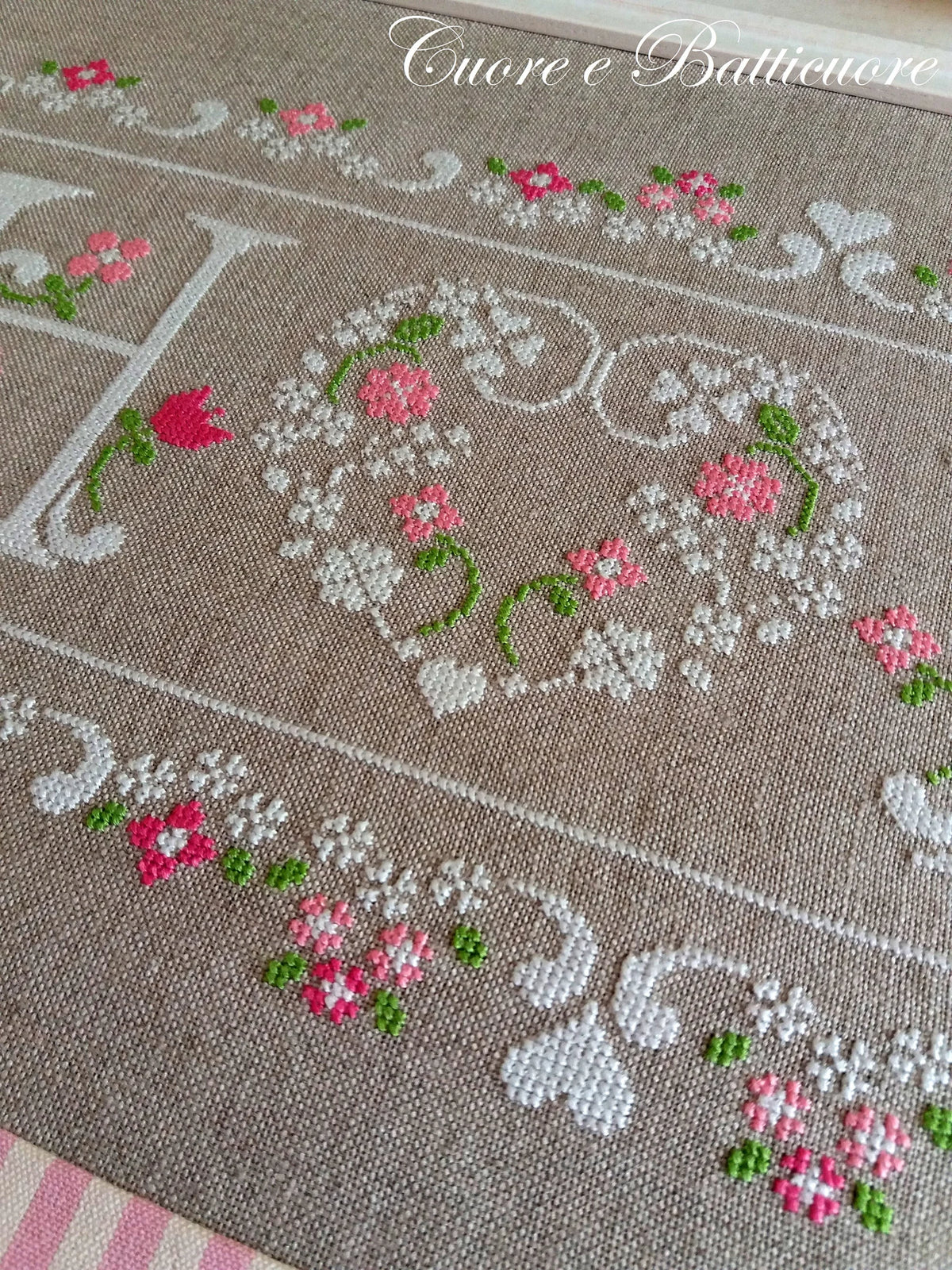 A close-up of a beige woven textile runner or rug featuring embroidered floral motifs in pink, white, and green, with decorative scrollwork and a large letter 'H' partially visible.
Photo by CuoreBatticuore.com [additional_image_link]
