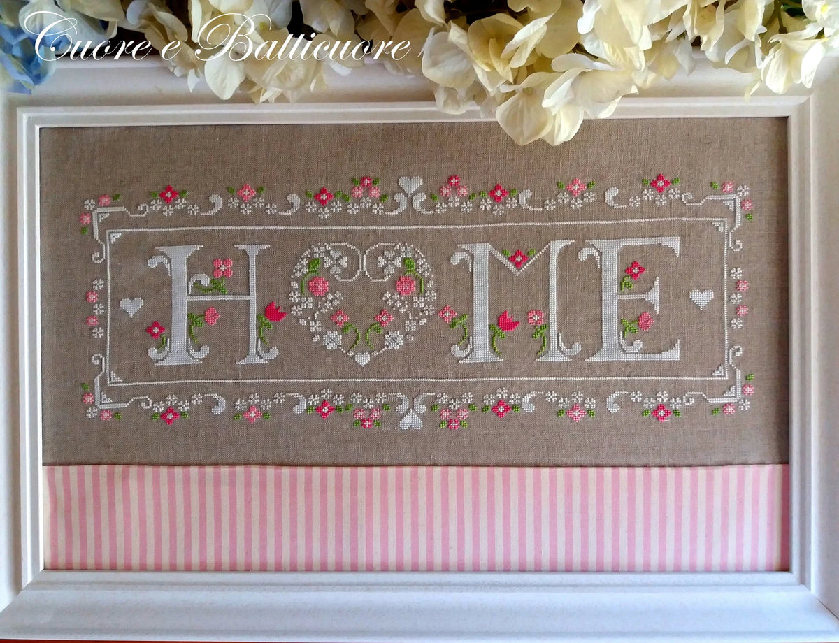 A framed cross-stitch piece featuring the word 'HOME' with a heart-shaped 'O' and floral borders, mounted on a brown linen background with a pink striped bottom panel. White flowers are arranged above the frame, and a watermark reading 'Cuore e Batticuore' appears in the upper left.
Photo by CuoreBatticuore.com [additional_image_link]