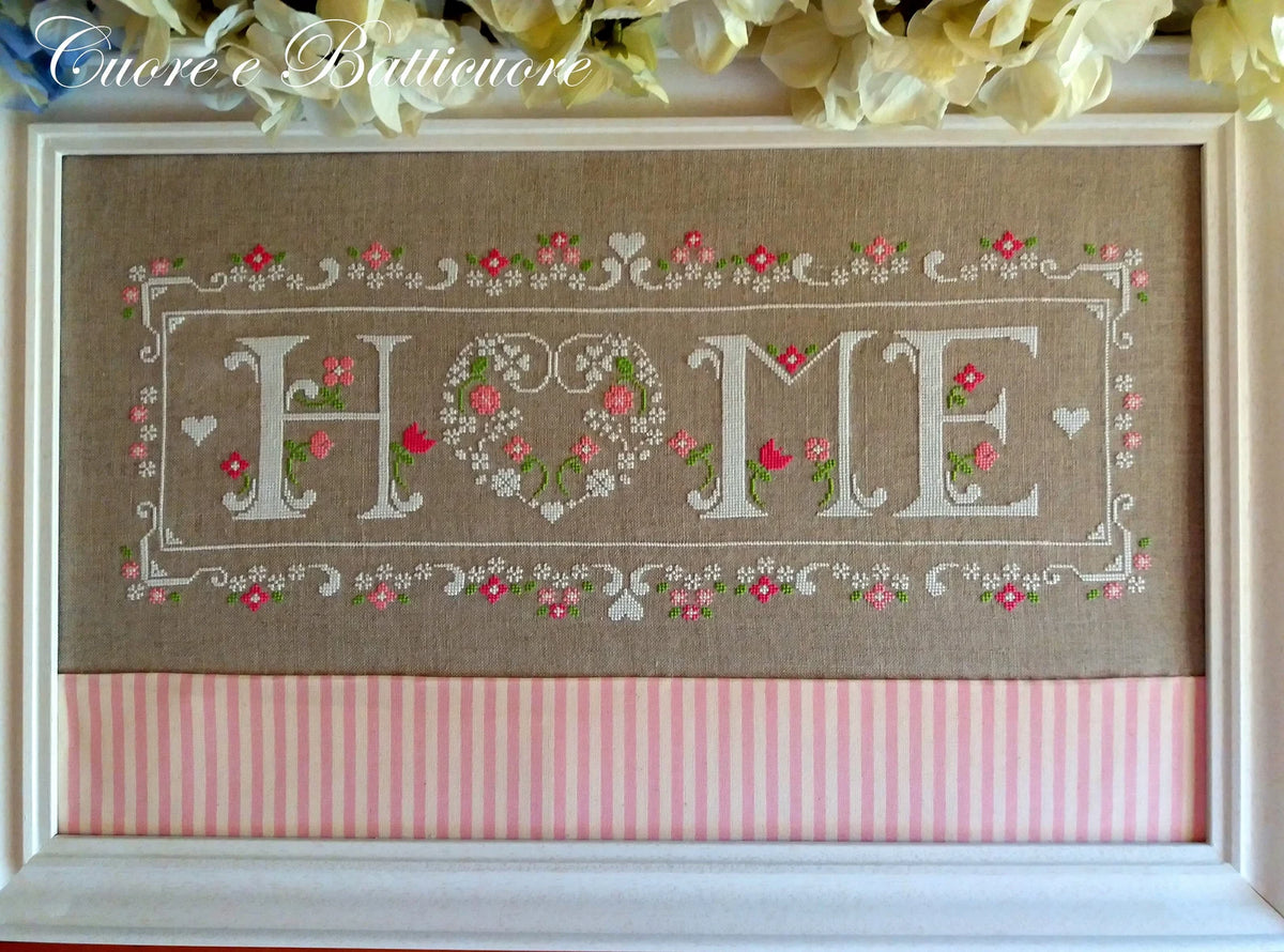 A framed decorative cross-stitch piece with the word “HOME” stitched in white, where the O is formed by floral hearts and leaves. The fabric background is tan, with pink and white striped fabric along the bottom and white flowers at the top of the frame.
Photo by CuoreBatticuore.com [additional_image_link]