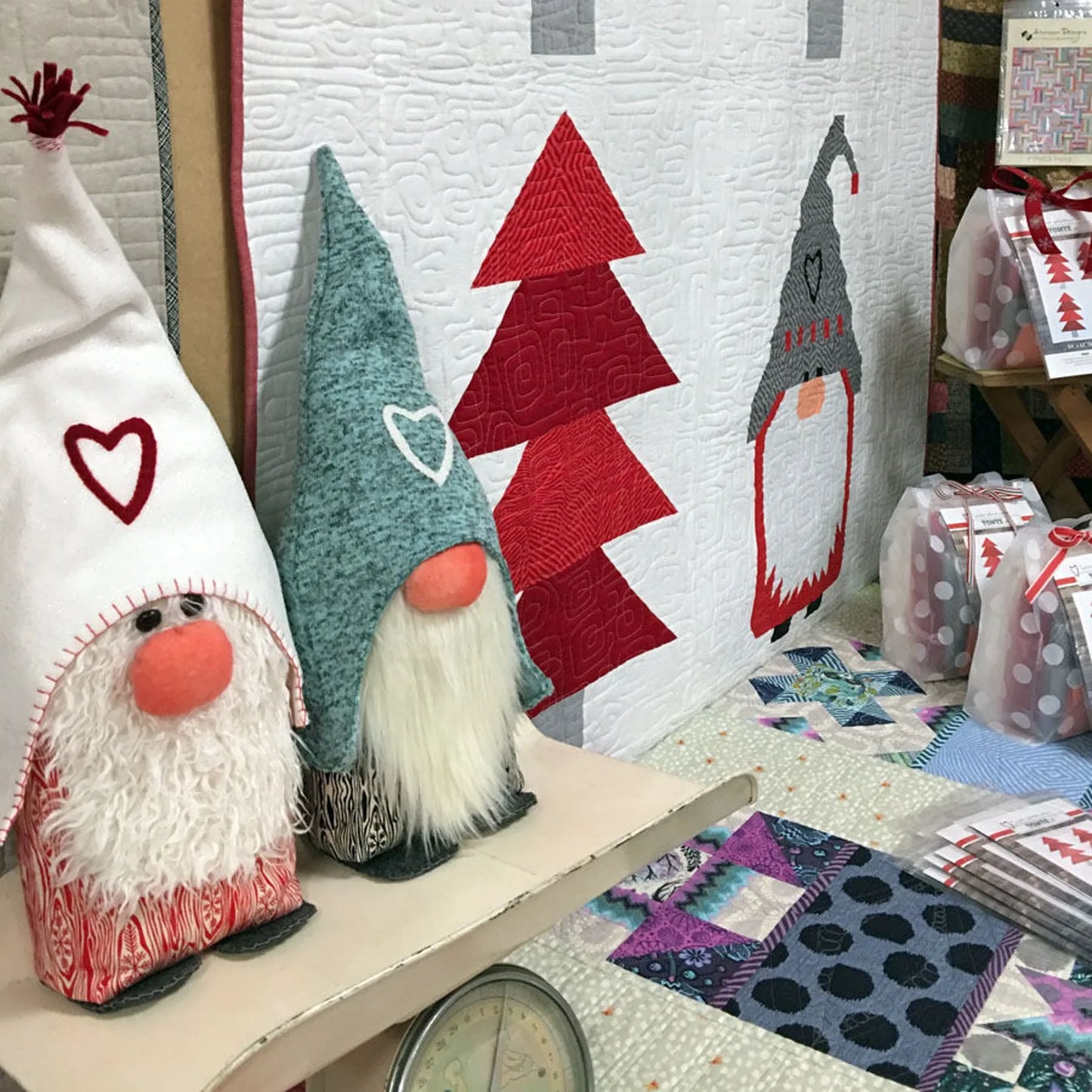Two quilted gnome decorations displayed on a table in a craft shop or studio setting, with a matching quilted wall hanging behind them showing gnomes and Christmas trees.
Photo by Sherri Noel [additional_image_link]