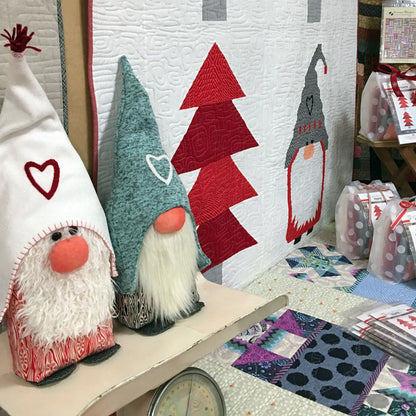 Two quilted gnome decorations displayed on a table in a craft shop or studio setting, with a matching quilted wall hanging behind them showing gnomes and Christmas trees.
Photo by Sherri Noel [additional_image_link]