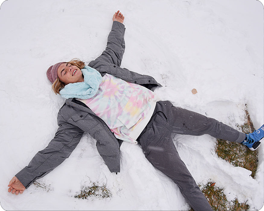 A person lying in the snow making a star angel face, wearing a gray puffer jacket, a pastel tie-dye shirt, a light blue scarf, and a pink knit hat.
Photo by Shannon Fabrics [additional_image_link]