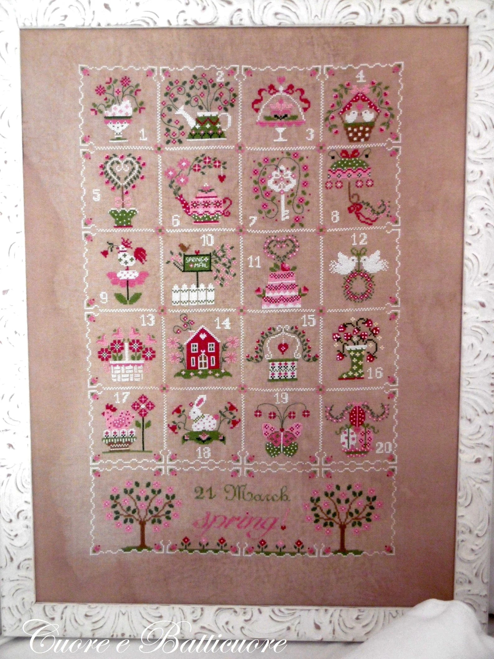 A square cross-stitch sampler titled '21 March Spring' featuring 21 numbered embroidered scenes of spring motifs like flowers, birds, a teapot, a house, and a rabbit, stitched on a dusty pink linen background and framed by a white lace mat.
Photo by CuoreBatticuore.com [image_link]