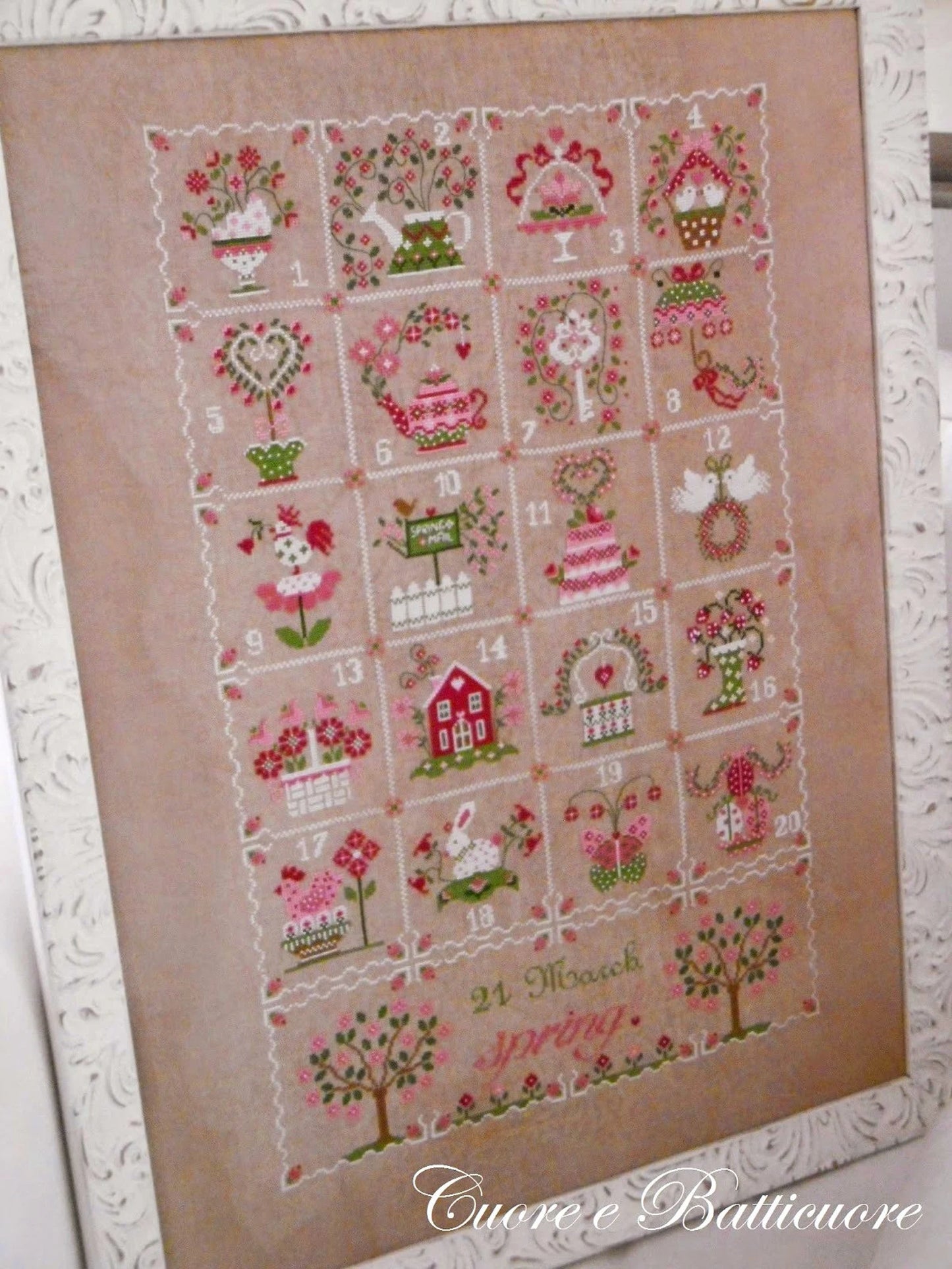 A framed cross-stitch piece featuring a spring-themed 24-day advent calendar with floral, garden, and house motifs on a beige linen background, shown against a white textured backdrop.
Photo by CuoreBatticuore.com [additional_image_link]