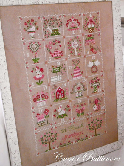 A framed cross-stitch piece featuring a spring-themed 24-day advent calendar with floral, garden, and house motifs on a beige linen background, shown against a white textured backdrop.
Photo by CuoreBatticuore.com [additional_image_link]
