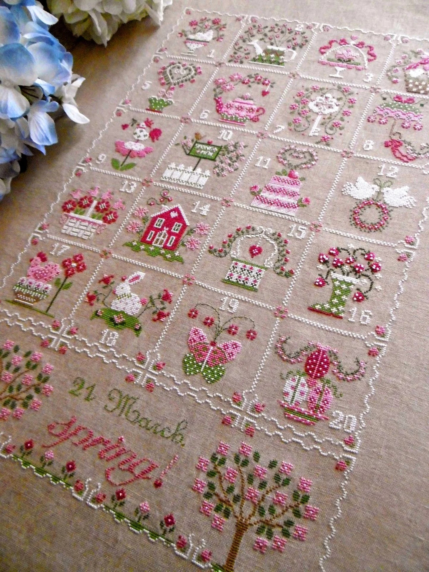 A square embroidered cross-stitch piece laid flat on a beige fabric background, featuring a grid of numbered scenes with pink flowers, green leaves, birds, houses, and the text '21 March Happy Birthday!' at the bottom.
Photo by CuoreBatticuore.com [additional_image_link]