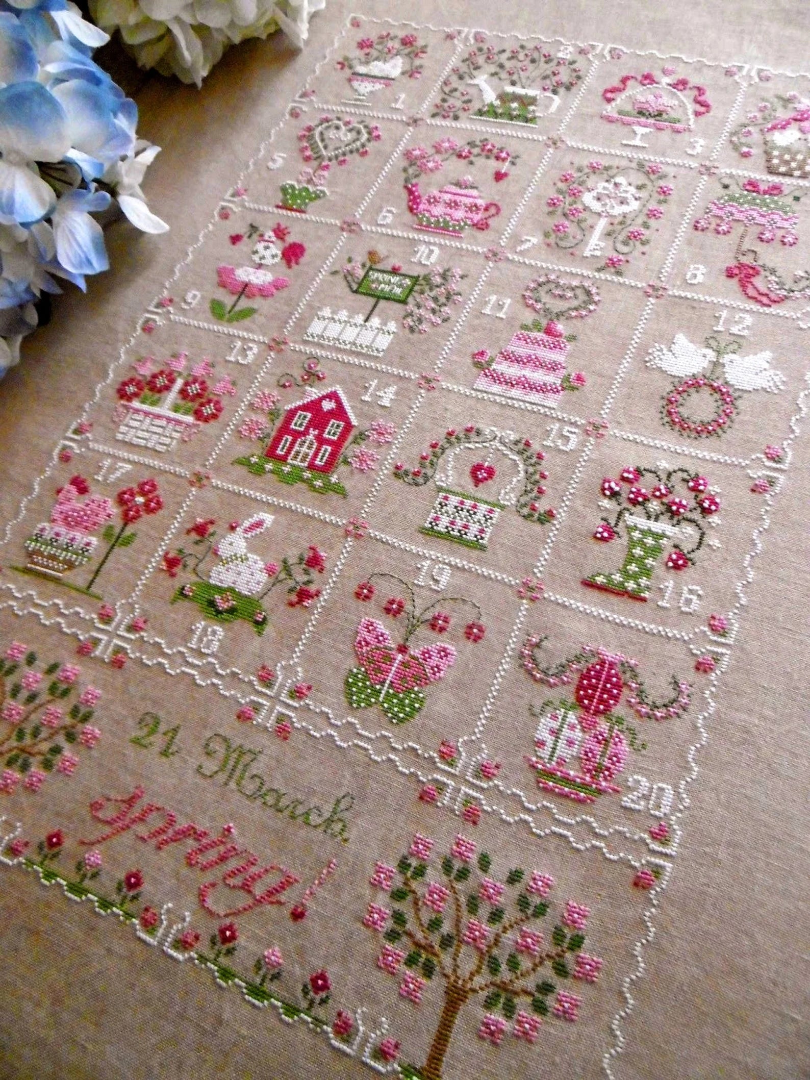 A square embroidered cross-stitch piece laid flat on a beige fabric background, featuring a grid of numbered scenes with pink flowers, green leaves, birds, houses, and the text '21 March Happy Birthday!' at the bottom.
Photo by CuoreBatticuore.com [additional_image_link]