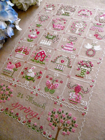 A square embroidered cross-stitch piece laid flat on a beige fabric background, featuring a grid of numbered scenes with pink flowers, green leaves, birds, houses, and the text '21 March Happy Birthday!' at the bottom.
Photo by CuoreBatticuore.com [additional_image_link]