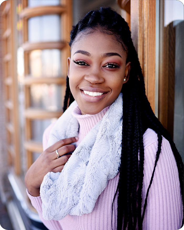 A smiling woman with long black braids wears a light pink ribbed turtleneck sweater and a fluffy pale blue-gray scarf, standing indoors near a wooden-framed window.
Photo by Shannon Fabrics [additional_image_link]