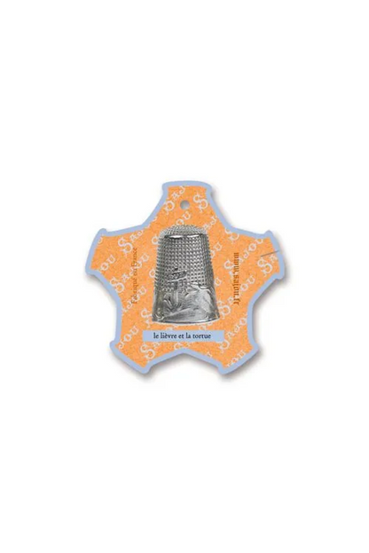 A small decorative thread card shaped like a star, featuring a silver thimble illustration in the center on a pale yellow and white background, placed on a white surface.
Photo by Sajou.fr [additional_image_link]
