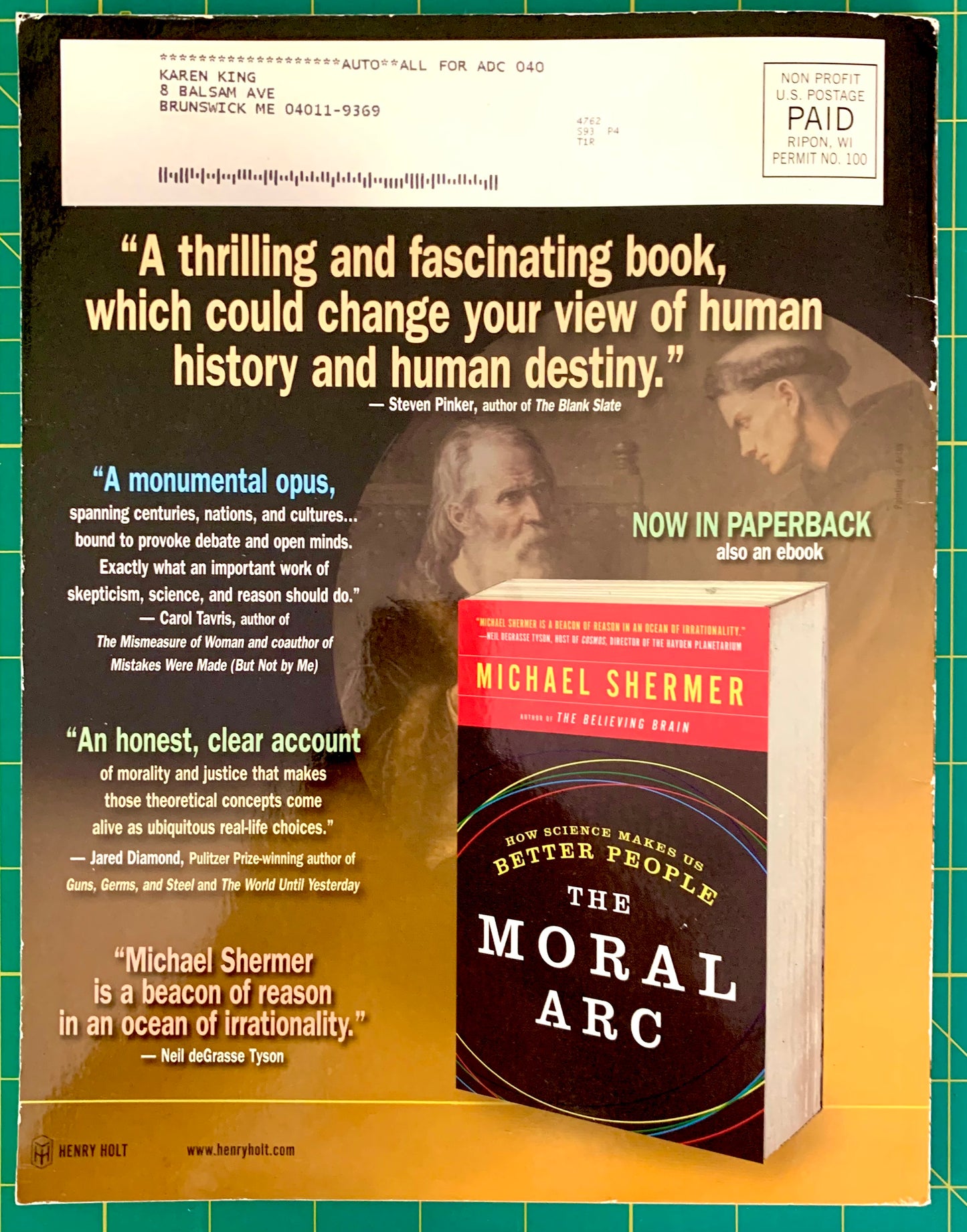 A vintage book advertisement for Michael Shermer's 'The Moral Arc' paperback, featuring glowing book cover artwork and several reader testimonials printed on a dark background, with a green cutting mat visible around the edges.
[additional_image_link]
