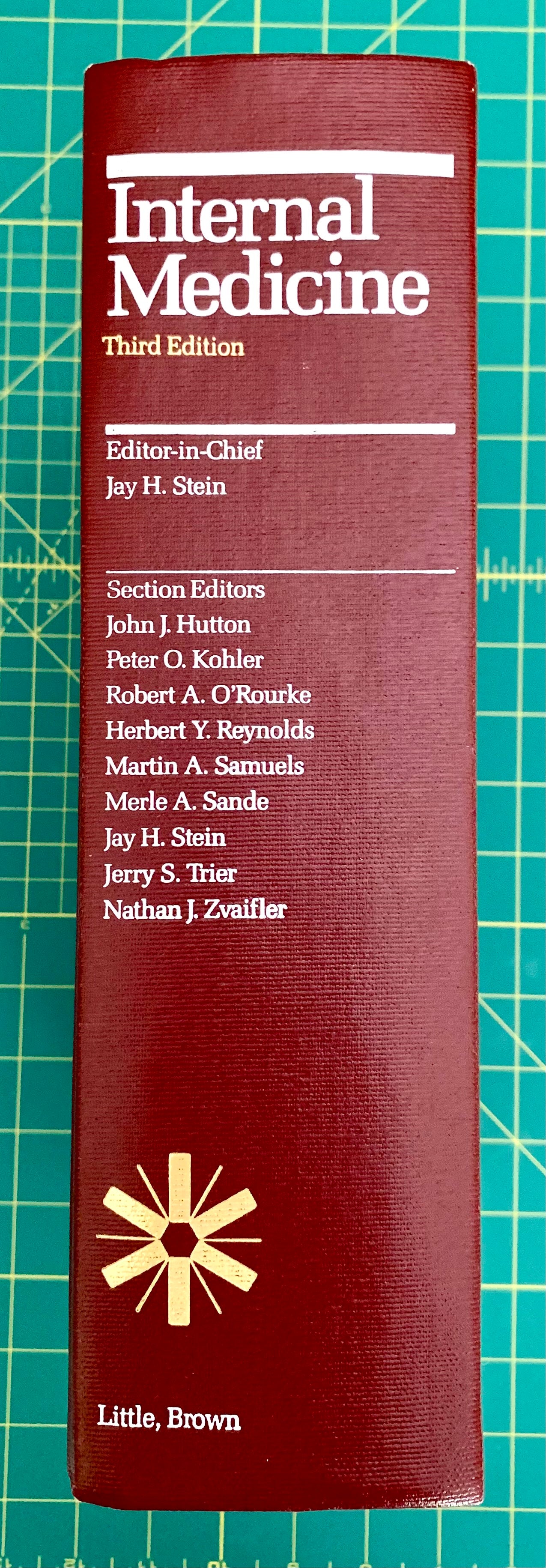 A hardcover book titled 'Internal Medicine, Third Edition' standing upright on a green cutting mat. The cover is dark red/burgundy with white text and a gold starburst logo near the bottom.
[additional_image_link]