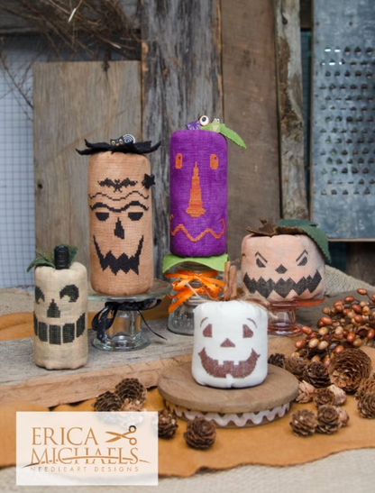 A collection of handmade burlap and fabric pumpkin lanterns decorated with black, orange, and purple Halloween motifs, arranged on a rustic wooden table with pinecones and a marshmallow pumpkin in front. A logo for 'Erica Michaels Needleart Designs' is visible in the bottom-left corner.
Photos by Erica Michaels [image_link]