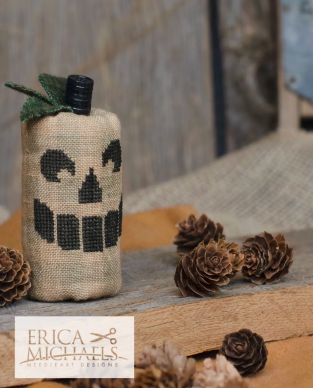 Toothy Jack.
A burlap-wrapped bottle styled as a pumpkin with a black stitched skull face and green leaves, set on a rustic wooden surface with pinecones. A white brand label for Erica Michaels Needleart Designs is visible in the lower left.
Photos by Erica Michaels [additional_image_link]