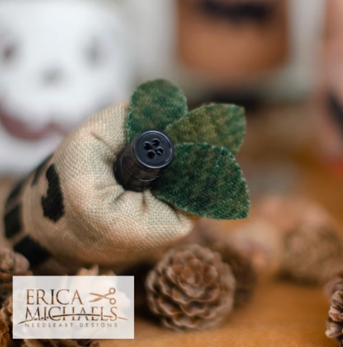 Toothy Jack top close up.
A close-up of a handmade Christmas stocking ornament by Erica Michaels Needleart Designs, featuring a beige burlap body with black knit stockings, green felt leaves, and a black button. The background is a warm, blurred setting with pinecones.
Photos by Erica Michaels [additional_image_link]