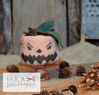 Stingy Jack.
A handmade fabric pumpkin needlepoint design on a small glass pedestal, decorated with green leaves and brown pinecones, with a rustic wooden background and a 'ERICA MICHAELS NEEDLEART DESIGNS' logo overlay in the bottom left.
Photos by Erica Michaels [additional_image_link]