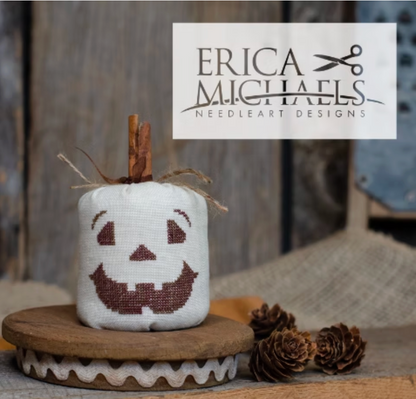 Classic Jack.
A small white fabric pumpkin-shaped needlepoint design with a brown stitched jack-o'-lantern face and a cinnamon stick stem, displayed on a wooden stand with pinecones. A white brand label reading Erica Michaels Needleart Designs is overlaid at the top right.
Photos by Erica Michaels [additional_image_link]