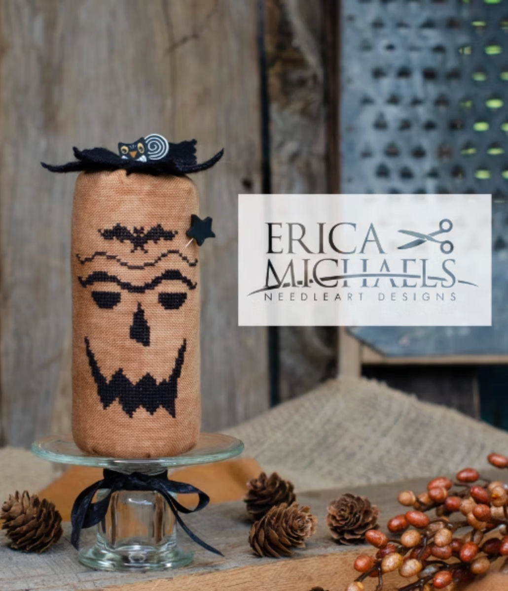 Scary Jack.
A needlepoint canvas featuring a black jack-o'-lantern face on a tan burlap-style background, displayed on a glass stand with pinecones and berries. A white brand label for Erica Michaels Needleart Designs is overlaid on the right side.
Photos by Erica Michaels [additional_image_link]