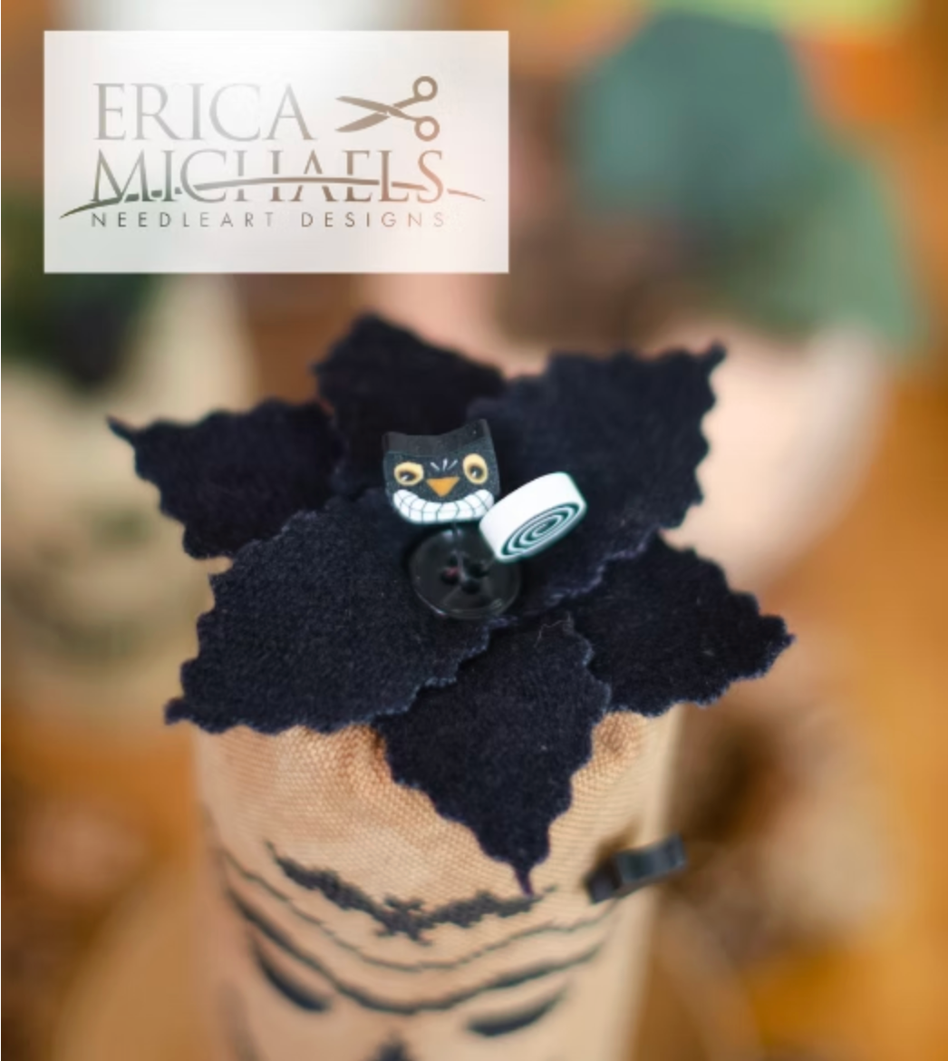 Scary Jack top close up.
A close-up of a needlework design featuring a black felt flower with a small owl embellishment and a button, mounted on a beige fabric-covered pin cushion. A white logo overlay for Erica Michaels Needleart Designs is visible in the top left corner.
Photos by Erica Michaels [additional_image_link]