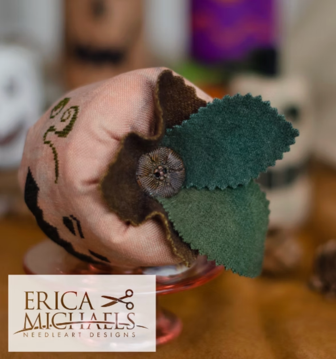 Stingy Jack top close up.
A handmade fabric needle case with a pink and brown floral design, a dark green felt leaf accent, and a central metallic button, displayed on a glass stand. A logo overlay for 'ERICA MICHAELS NEEDLEART DESIGNS' is visible in the bottom left corner.