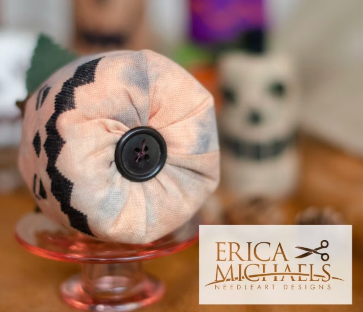 Stingy Jack bottom close up.
A handmade fabric needlecase with a black button on a pink glass stand, featuring a tie-dye pattern of pink, gray, and white with black stitched accents. A logo for 'ERICA MICHAELS NEEDLEART DESIGNS' is visible in the bottom right corner.