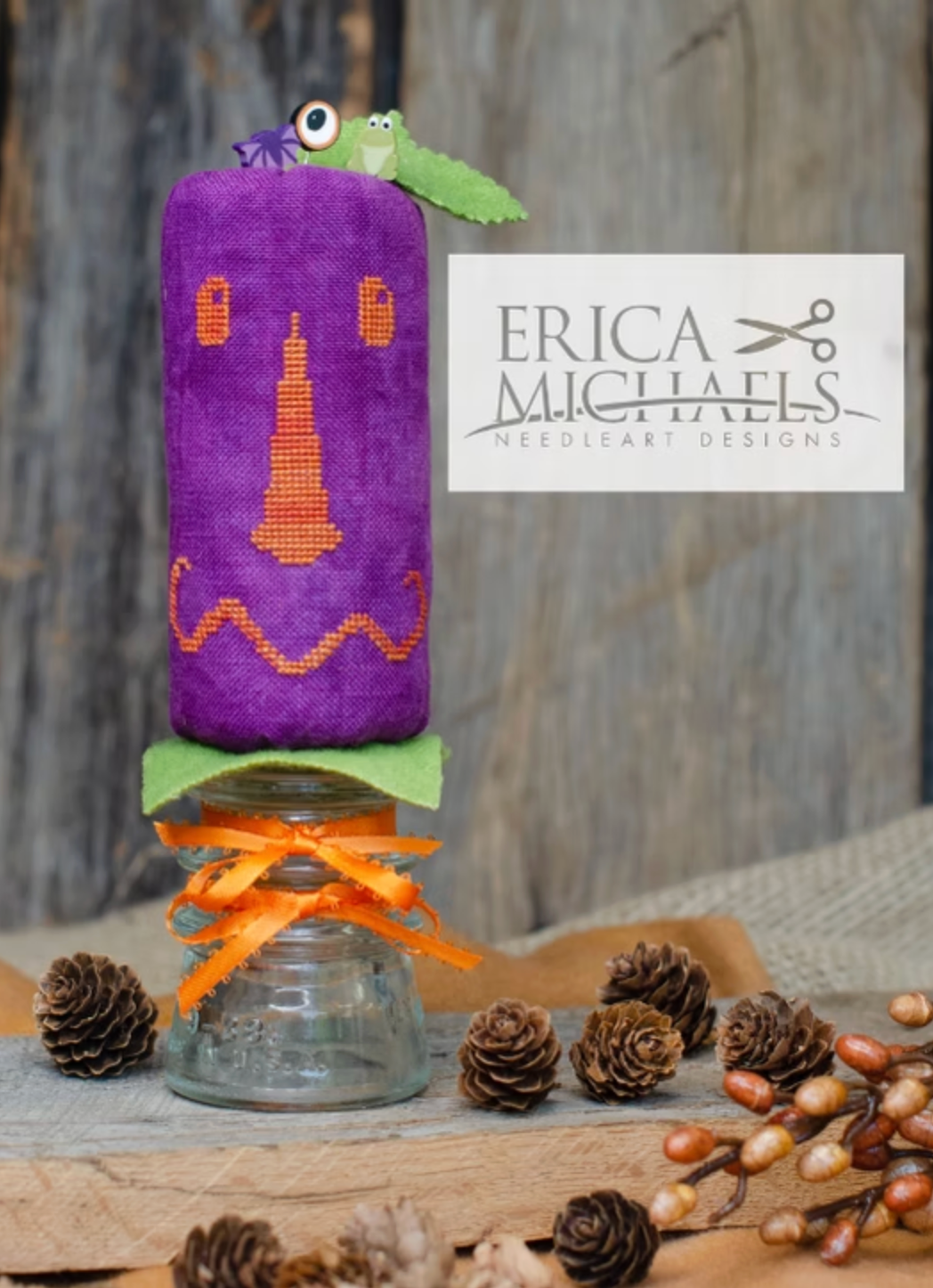 Skinny Jack.
A purple fabric-covered needlepoint canvas with an orange jack-o'-lantern face and green leaf accents, displayed on a glass jar with an orange ribbon, surrounded by pinecones and a rustic wood background. A white label reads Erica Michaels Needleart Designs.
Photos by Erica Michaels [additional_image_link]