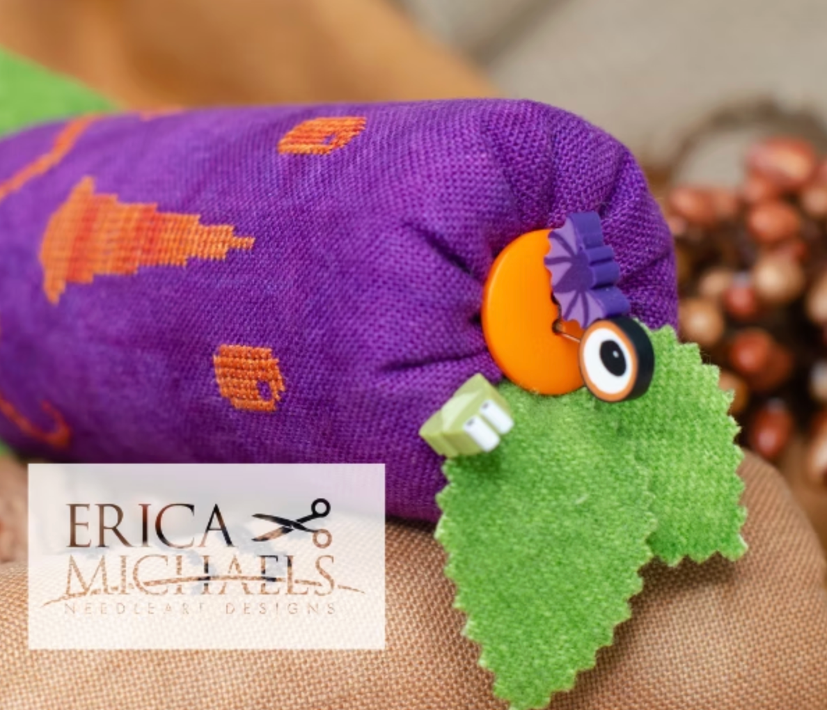 Skinny Jack top close up.
A close-up of a handmade fabric pouch in purple with orange embroidered motifs, featuring a green leaf-shaped charm with a button eye and purple flower accent, resting on a beige textured surface. A white brand label for Erica Michaels Needlecraft Designs is overlaid in the lower left.
Photos by Erica Michaels [additional_image_link]